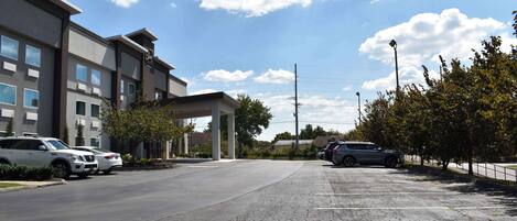 Un parking avec plusieurs voitures, un bâtiment doté d’une entrée abritée et des arbres.