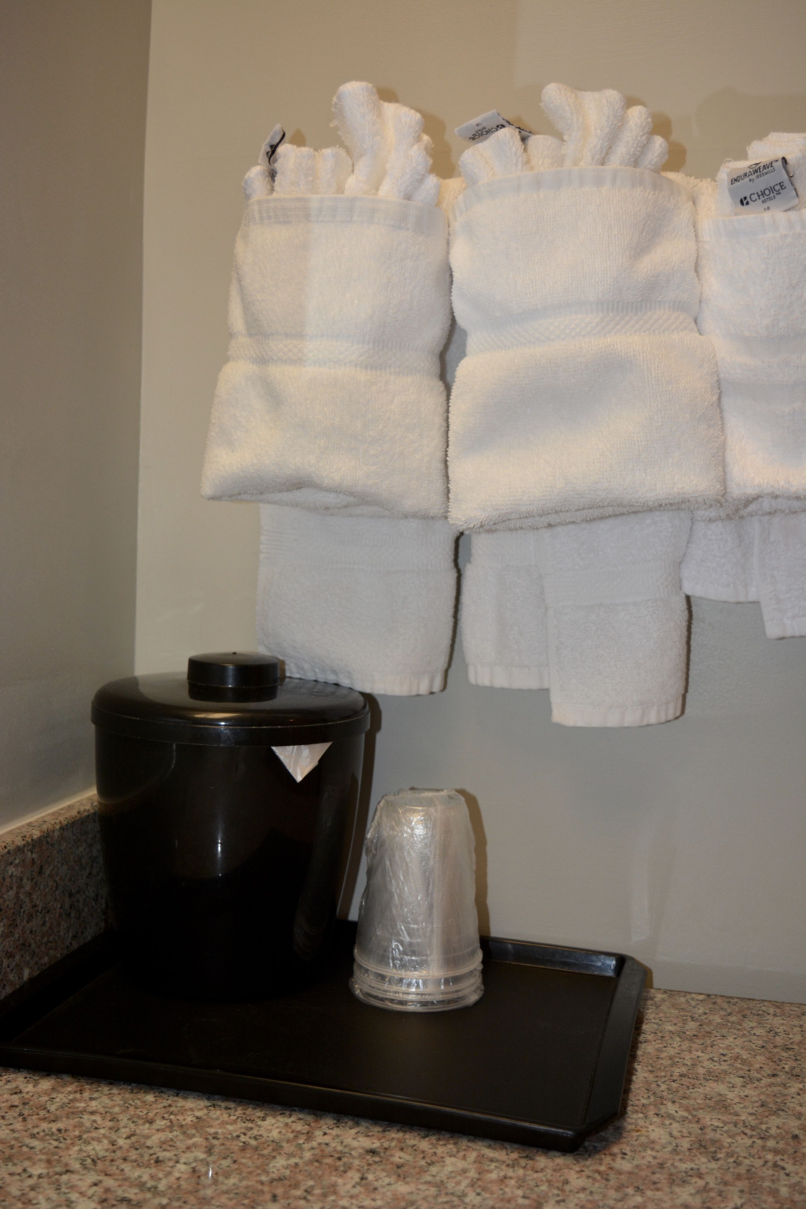 A hotel bathroom with a towel rack holding several white towels, a black coffee maker, and a glass carafe on a tray.