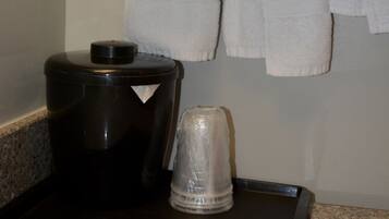 A hotel bathroom with a towel rack holding several white towels, a black coffee maker, and a glass carafe on a tray.