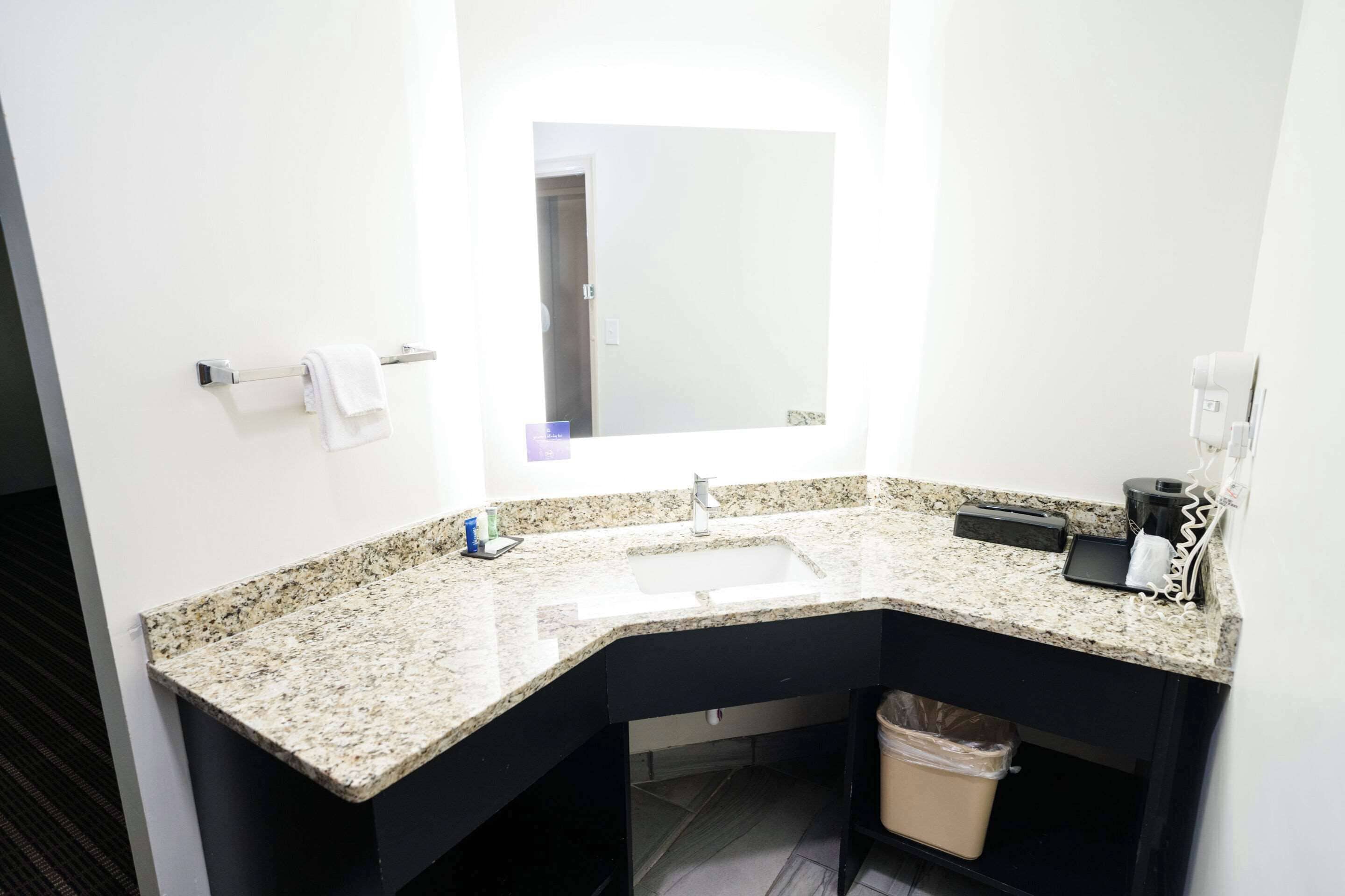 Un baño moderno con encimera de granito, un lavamanos, un espejo, un perchero, un cesto de basura y una cafetera.