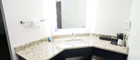 Un baño moderno con encimera de granito, un lavamanos, un espejo, un perchero, un cesto de basura y una cafetera.