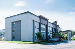 A modern multi-story building with a flat roof, large windows, and a parking area with cars.