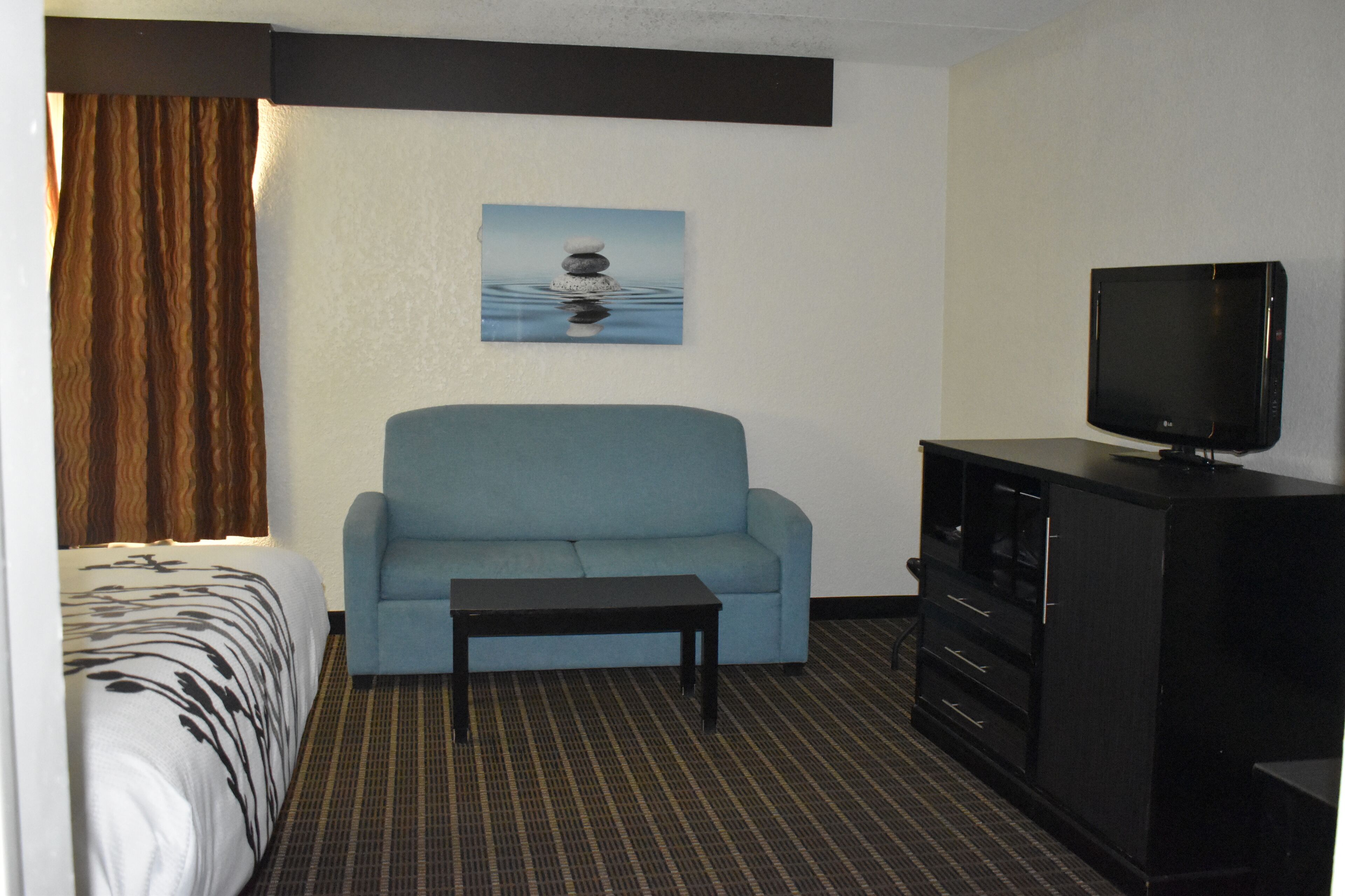 Una habitación de hotel con una cama, un sofá azul, una mesita, un televisor sobre un mueble y un cuadro en la pared.