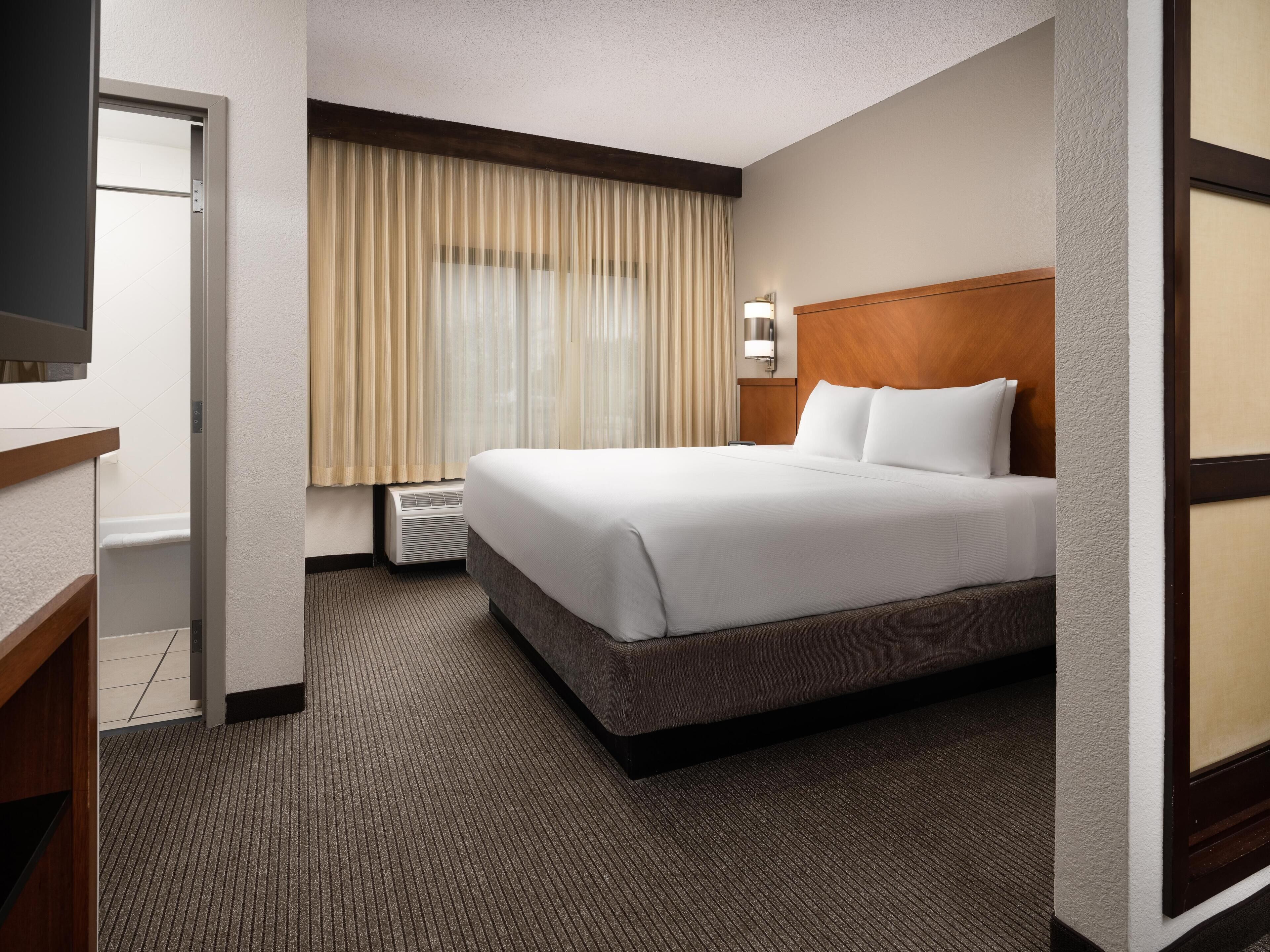 A hotel room with a bed, a desk, a window with curtains, a wall-mounted lamp, and a carpeted floor.