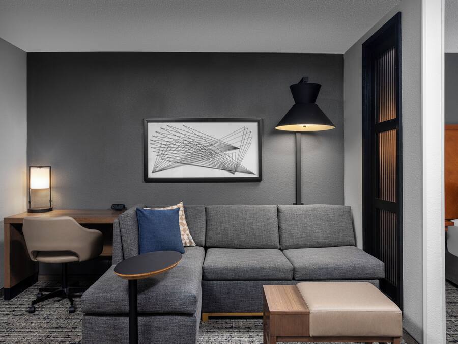 Et moderne hotelværelse med en grå sofa, et lille rundt bord, et skrivebord med stol, en sort gulvlampe og et indrammet abstrakt kunstværk på væggen.