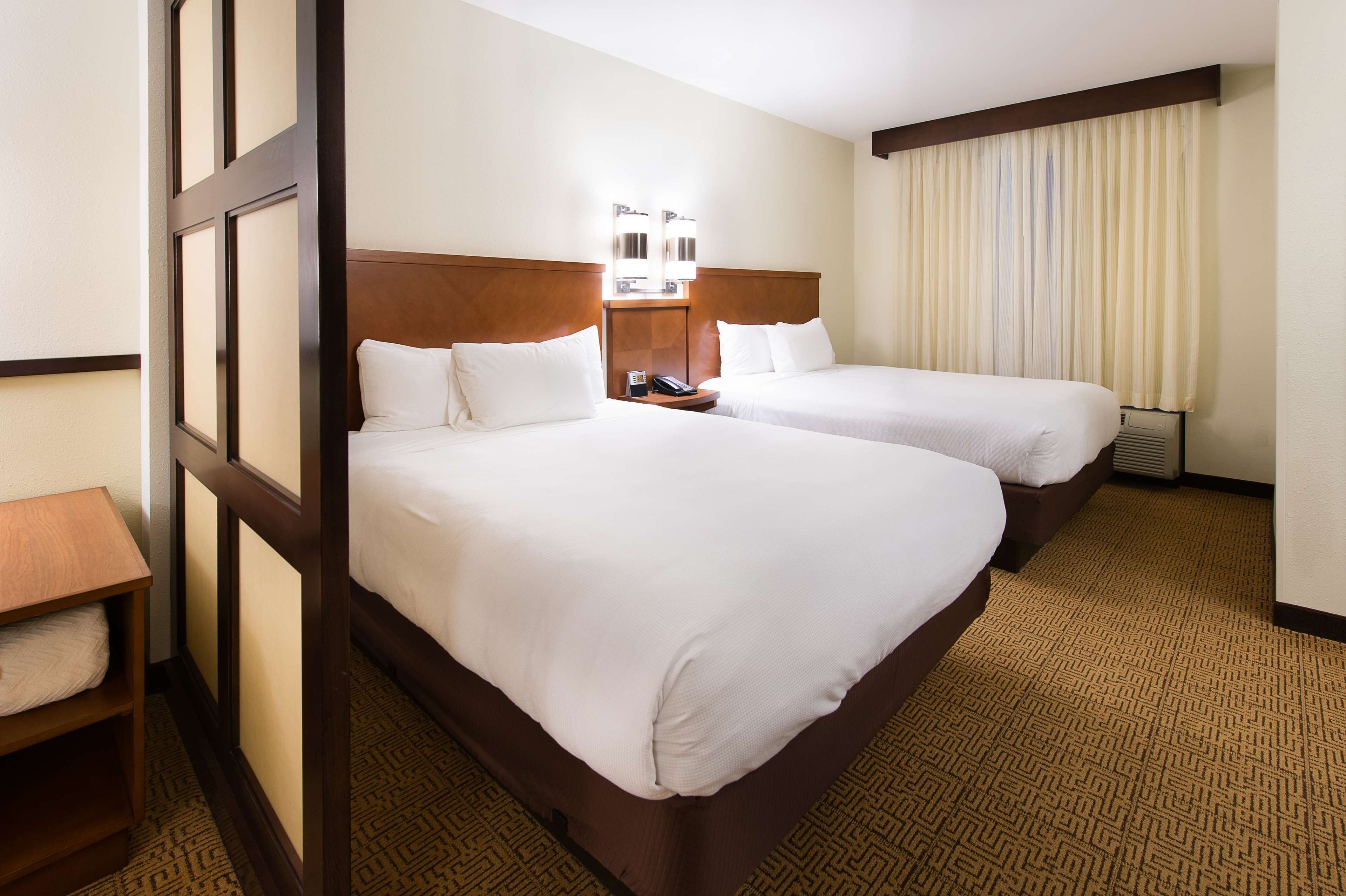 A hotel room with two beds, a wooden headboard, a bedside table with a lamp, and a large window with curtains.