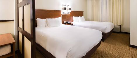A hotel room with two beds, a wooden headboard, a bedside table with a lamp, and a large window with curtains.