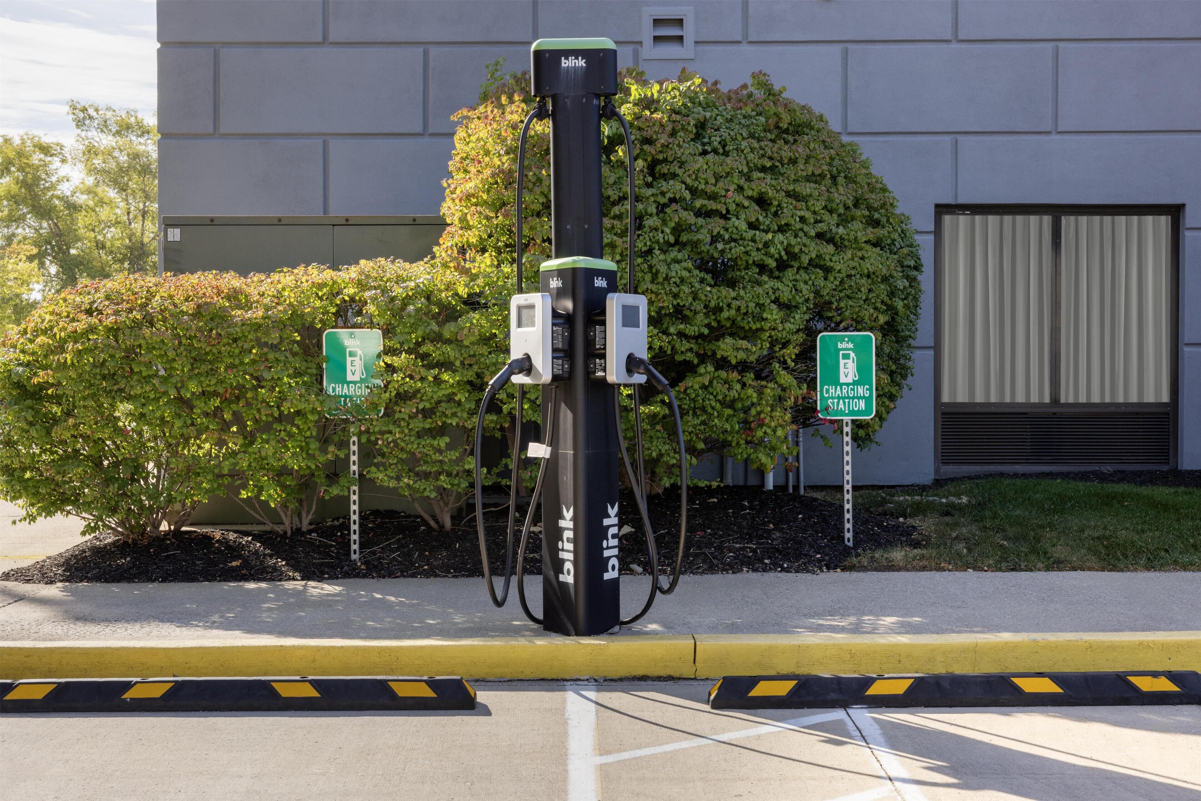 Estación de carga para vehículos eléctricos Blink con dos conectores, situada en un estacionamiento con un edificio al fondo.