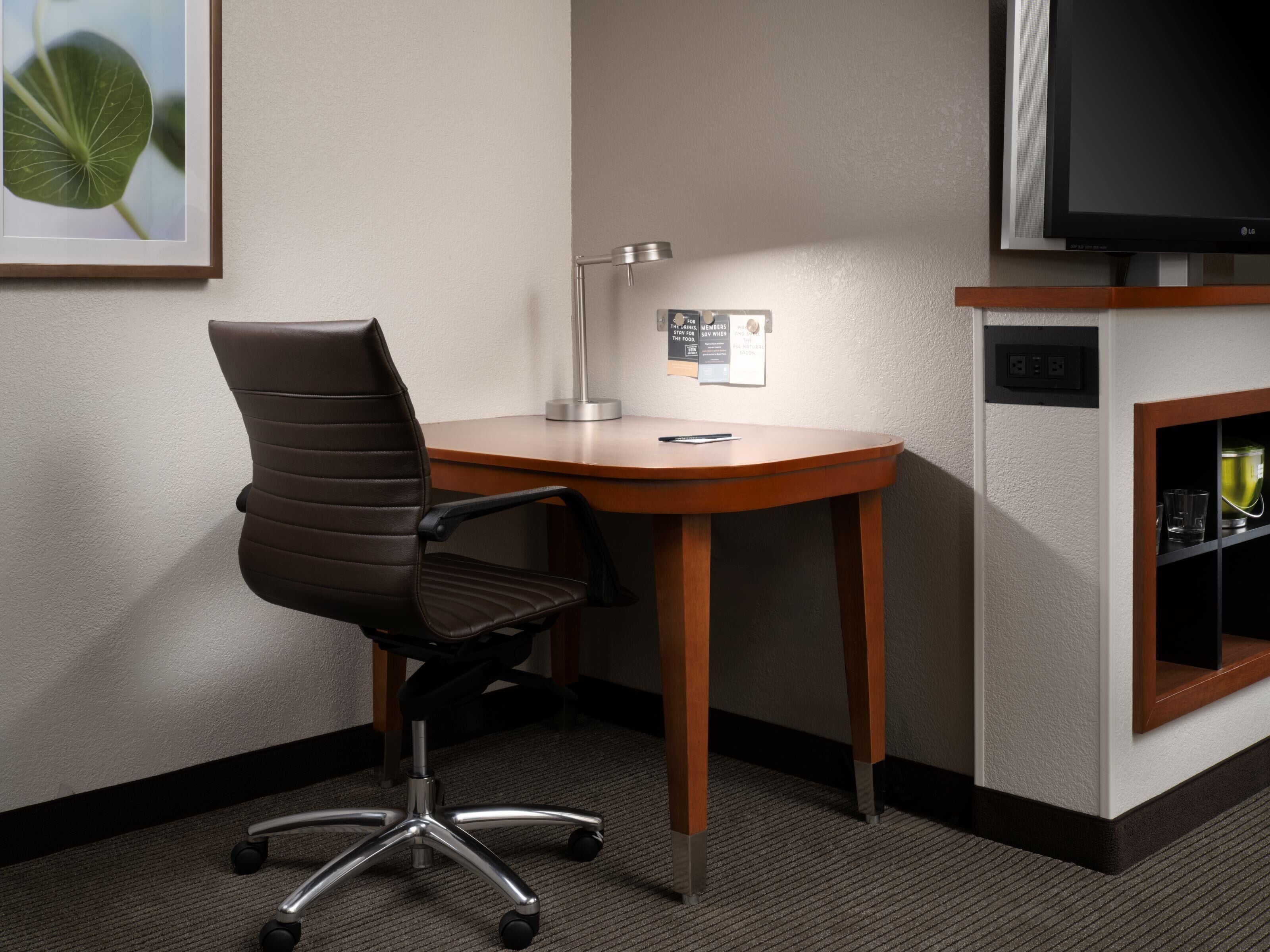 A hotel room with a desk, chair, lamp, TV, and a shelf with items.