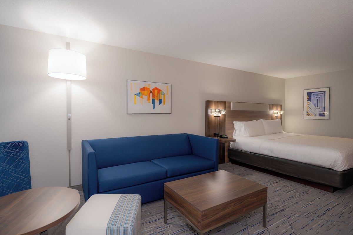 A hotel room with a blue sofa, a wooden coffee table, a bed with white bedding, and two wall-mounted lamps.
