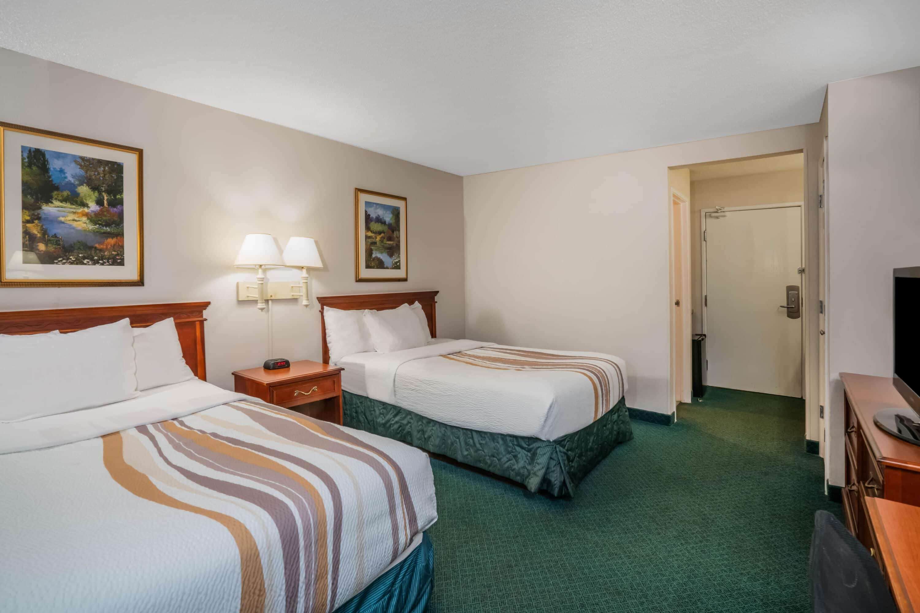A hotel room with two beds, a desk, a chair, a TV, and two paintings on the wall.