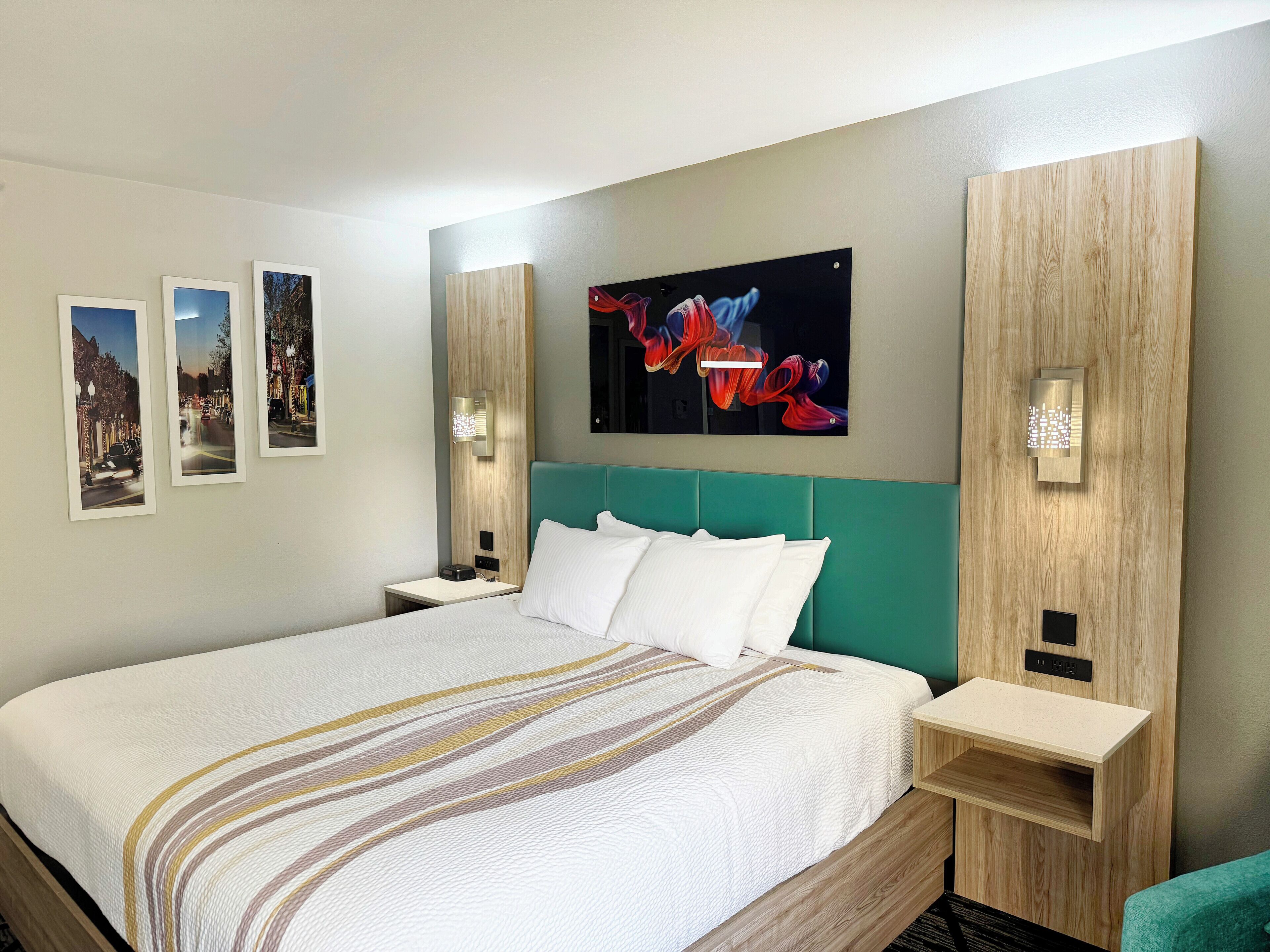 A hotel room with a large bed, a headboard, a nightstand, a wall-mounted light, and three framed pictures.