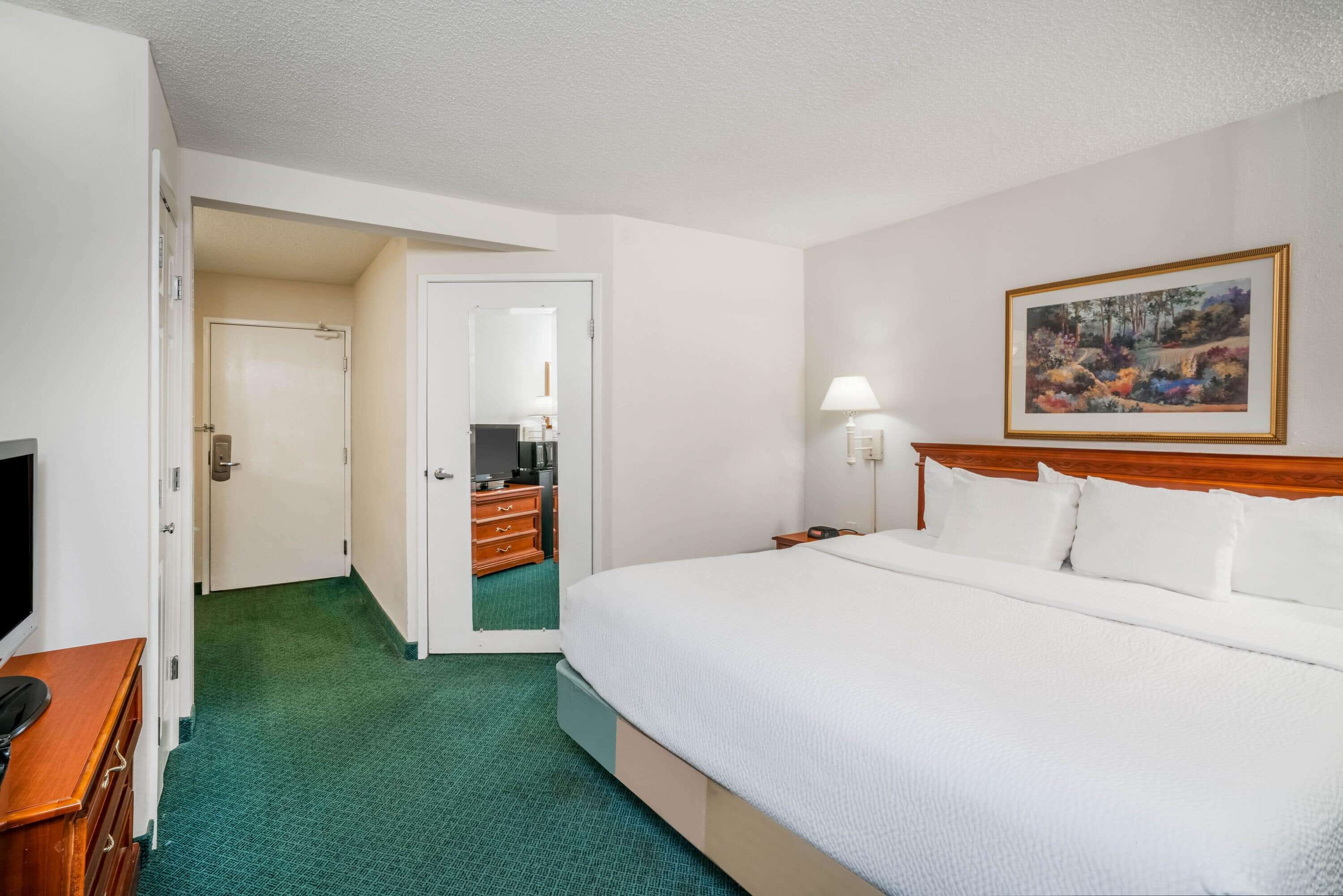 A hotel room with a large bed, a wooden headboard, a painting on the wall, a desk with a lamp, and a green carpet.