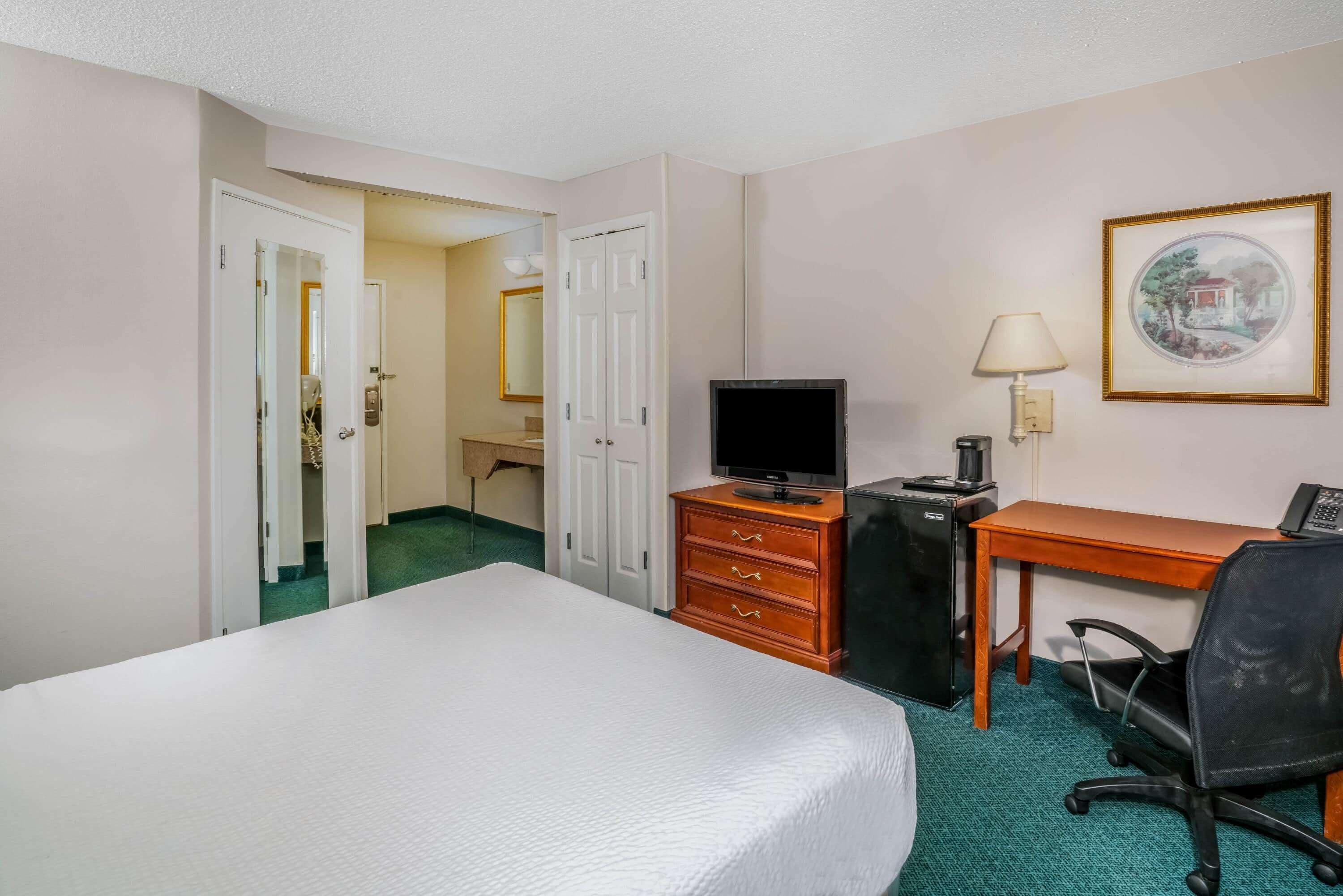 A hotel room with a bed, a desk, a chair, a television, and a desk lamp.