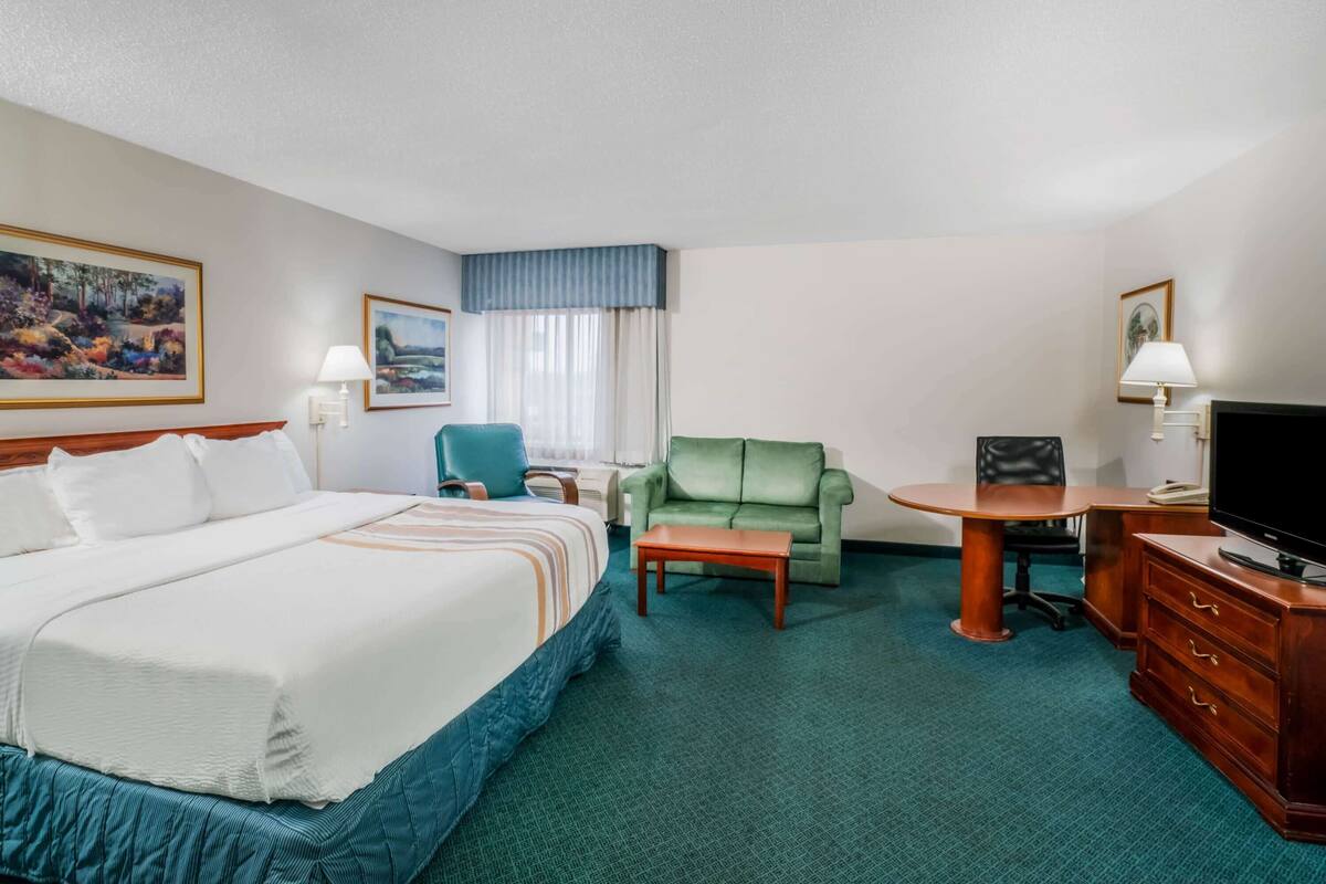 Una habitació d'hotel amb un llit gran, un otomà verd, una taula petita, un televisor i un escriptori amb calaixos.
