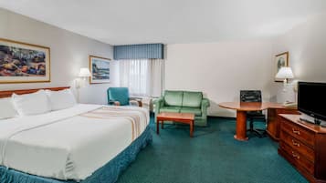Quarto de hotel com uma cama grande, uma poltrona verde, uma mesa pequena, uma televisão e uma escrivaninha com gavetas.