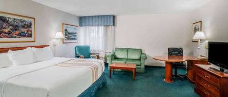 Quarto de hotel com uma cama grande, uma poltrona verde, uma mesa pequena, uma televisão e uma escrivaninha com gavetas.