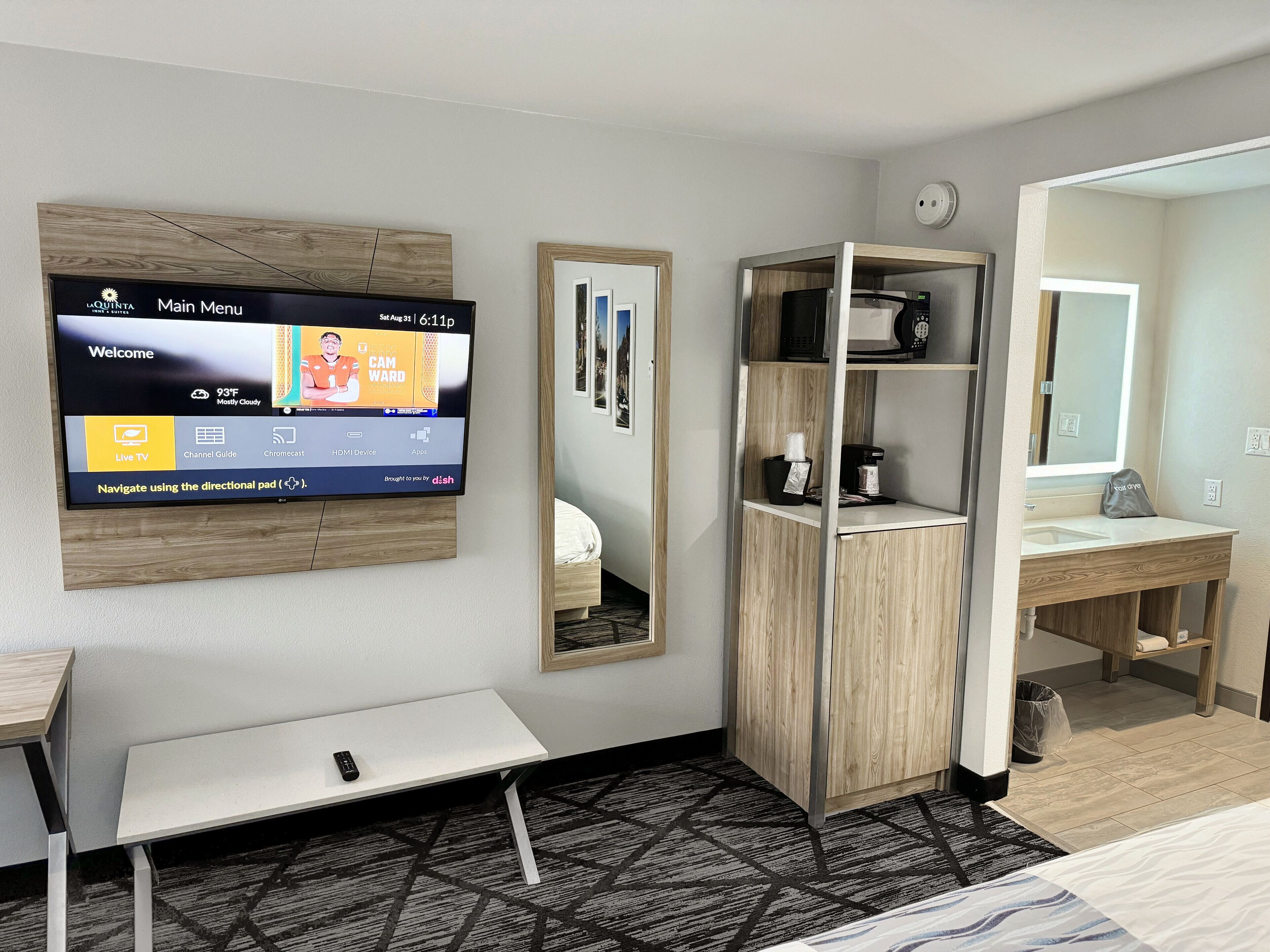A hotel room with a mounted TV displaying the main menu, a bedside table with a remote control, a mirror, a wardrobe, and a bathroom with a sink and mirror.