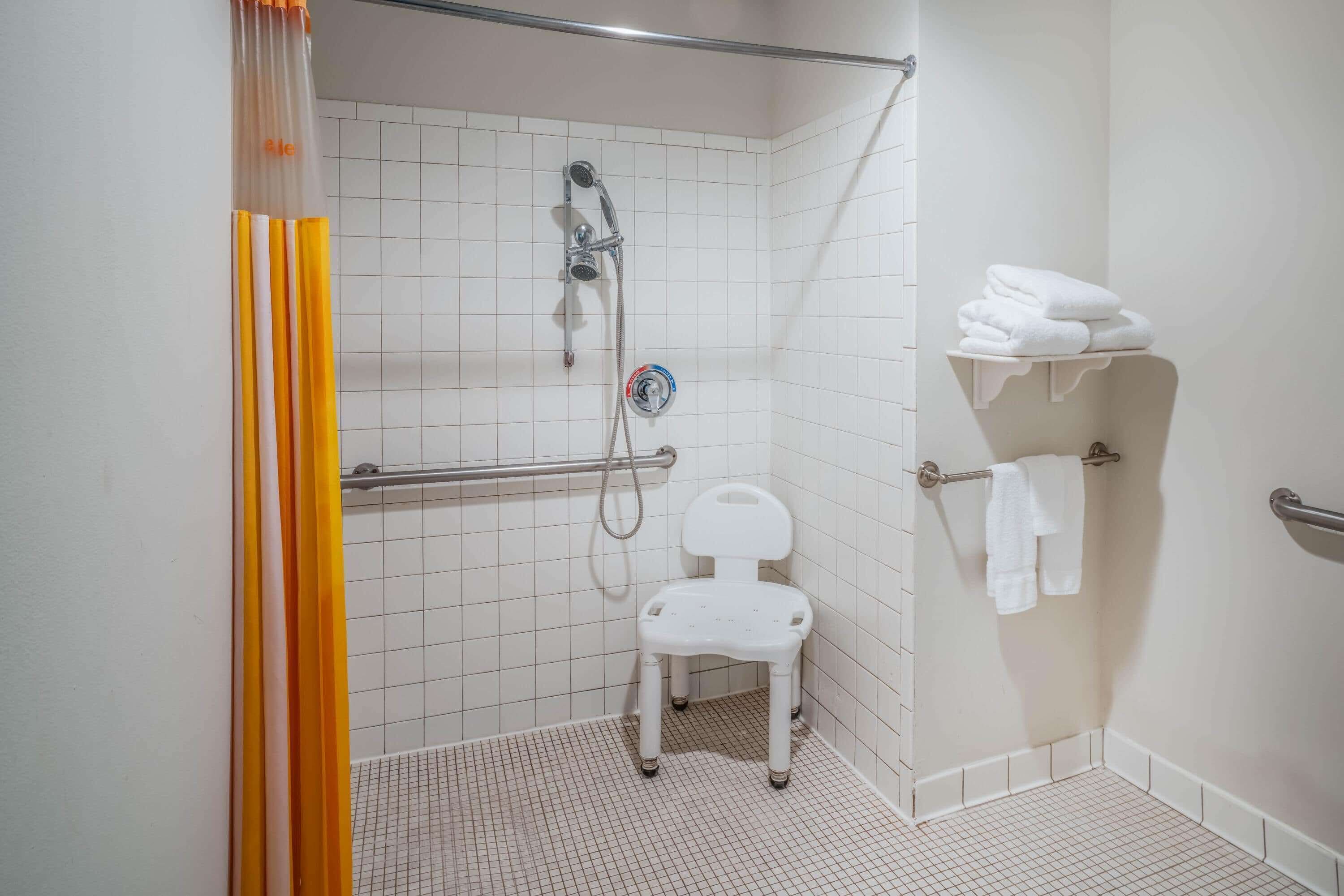 A small, white, tiled bathroom with a shower, a white chair, and a towel rack.