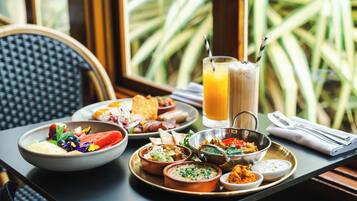 Un bufé de desayuno con diversos platos, un vaso de zumo de naranja y vistas a palmeras a través de una ventana.