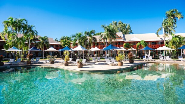 A resort pool area with lounge chairs and umbrellas, surrounded by palm trees and a building with a red and white facade. - Cable Beach Club Resort & Spa (Cable Beach)