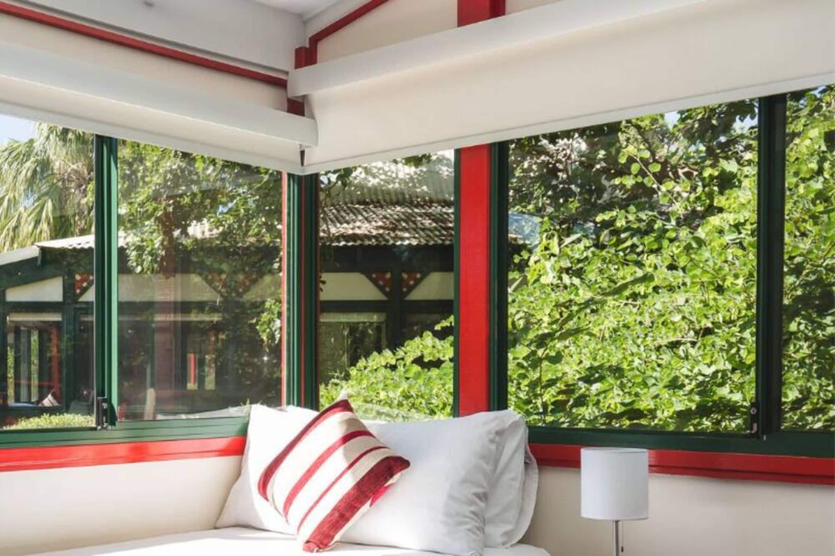 A room with a bed, a window with red trim, and a view of greenery outside.