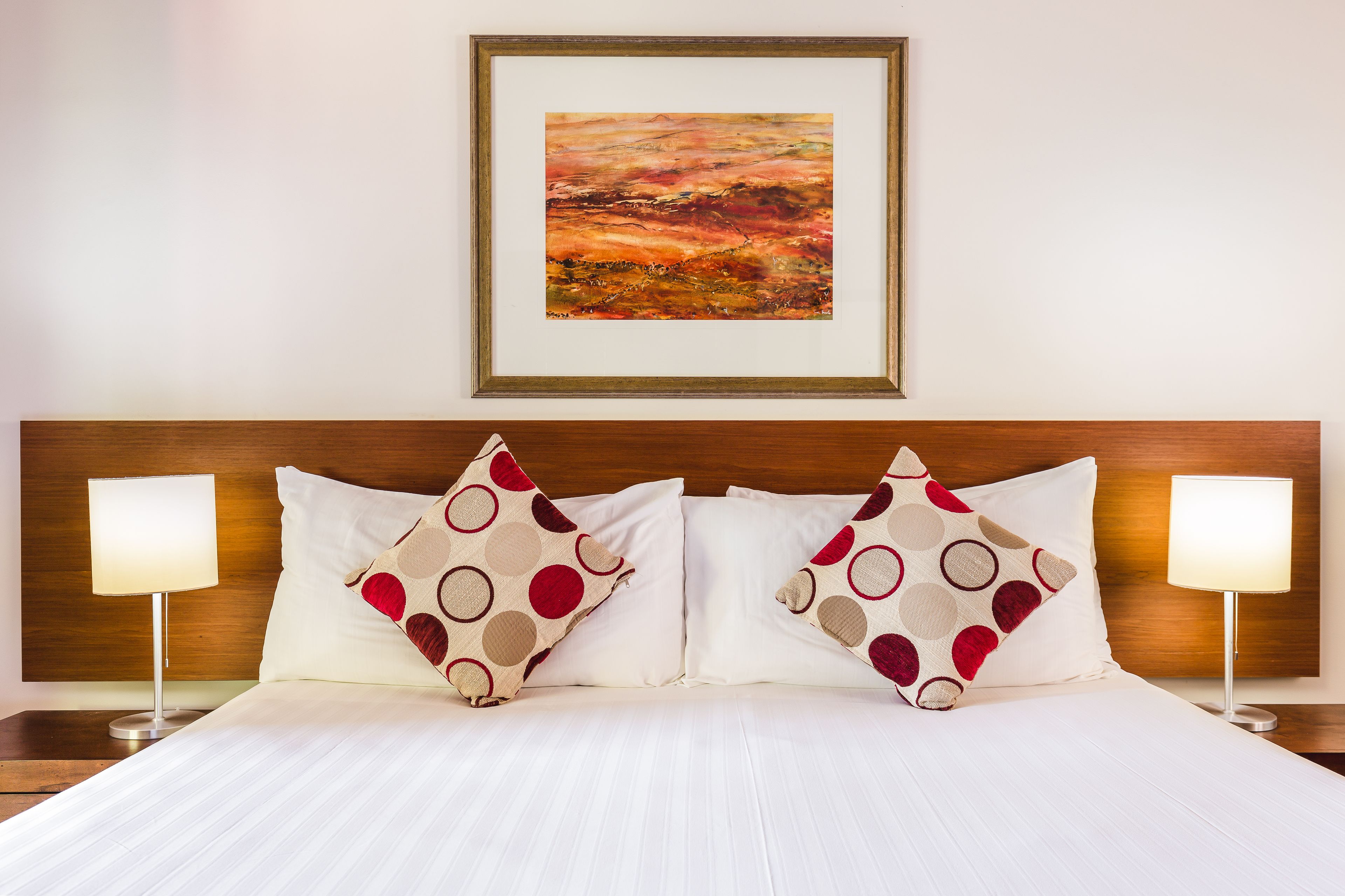 Cama bem arrumada com roupa de cama branca e duas almofadas decorativas com padrão em vermelho e bege. Há um quadro emoldurado acima da cama e duas luminárias de cabeceira, uma de cada lado.