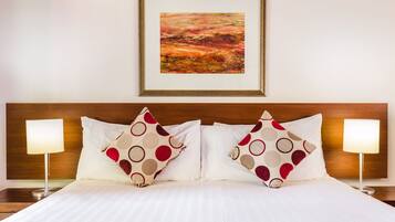 Cama bem arrumada com roupa de cama branca e duas almofadas decorativas com padrão em vermelho e bege. Há um quadro emoldurado acima da cama e duas luminárias de cabeceira, uma de cada lado.