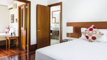 Une chambre d’hôtel avec un plancher en bois, un lit avec une literie blanche et des oreillers à motifs, une table de chevet avec une lampe, et une salle de bain visible à travers une porte ouverte.