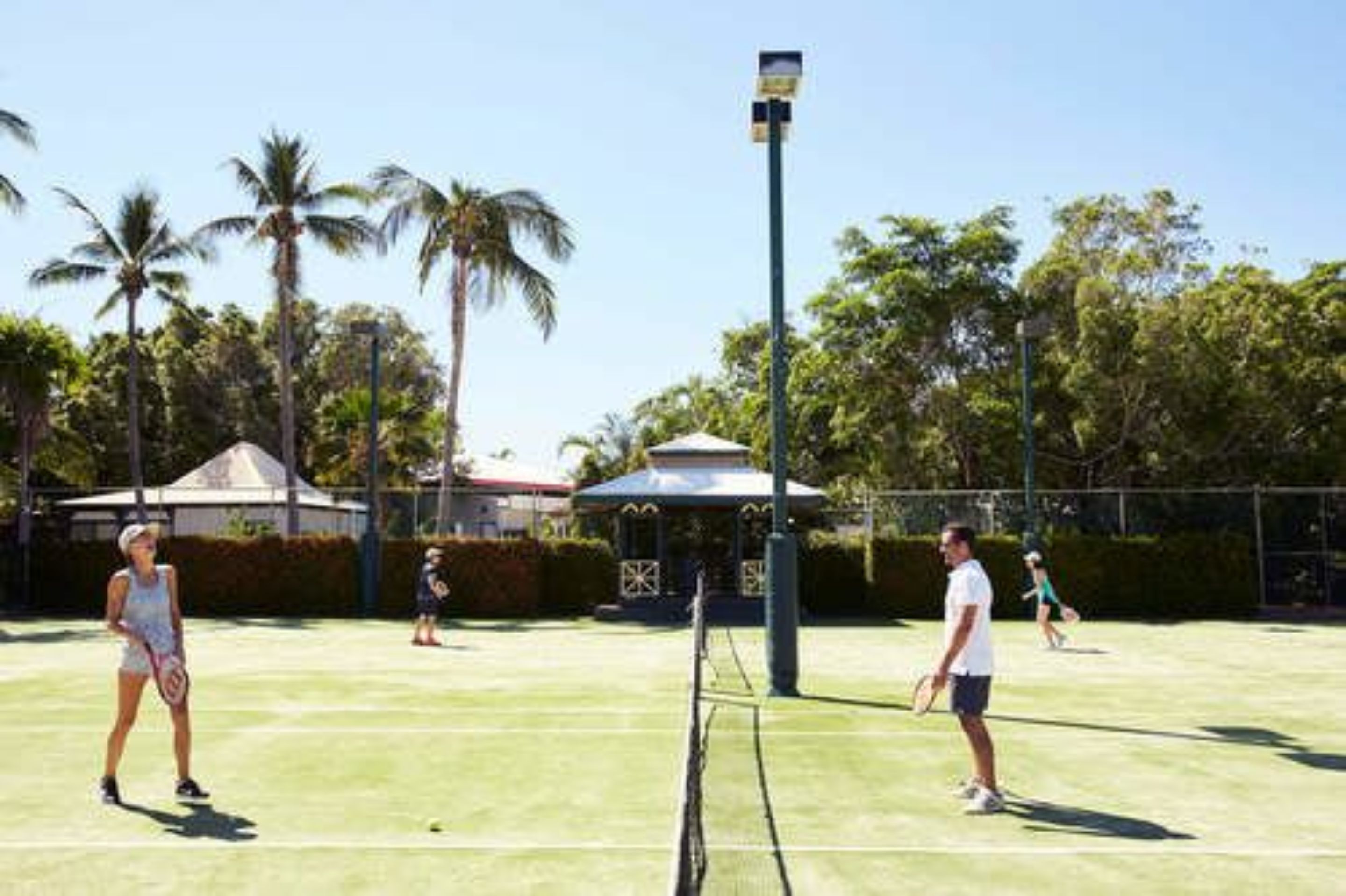A tennis court with players, palm trees, and a gazebo in the background.