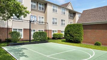An outdoor basketball court surrounded by a brick building with windows and a green lawn.