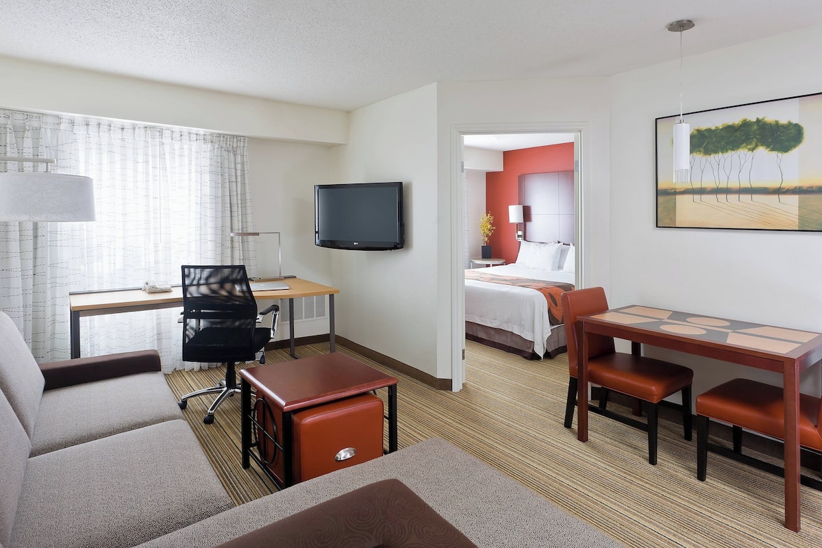 A hotel room with a bed, a desk with a chair, a TV, and a dining area with chairs.