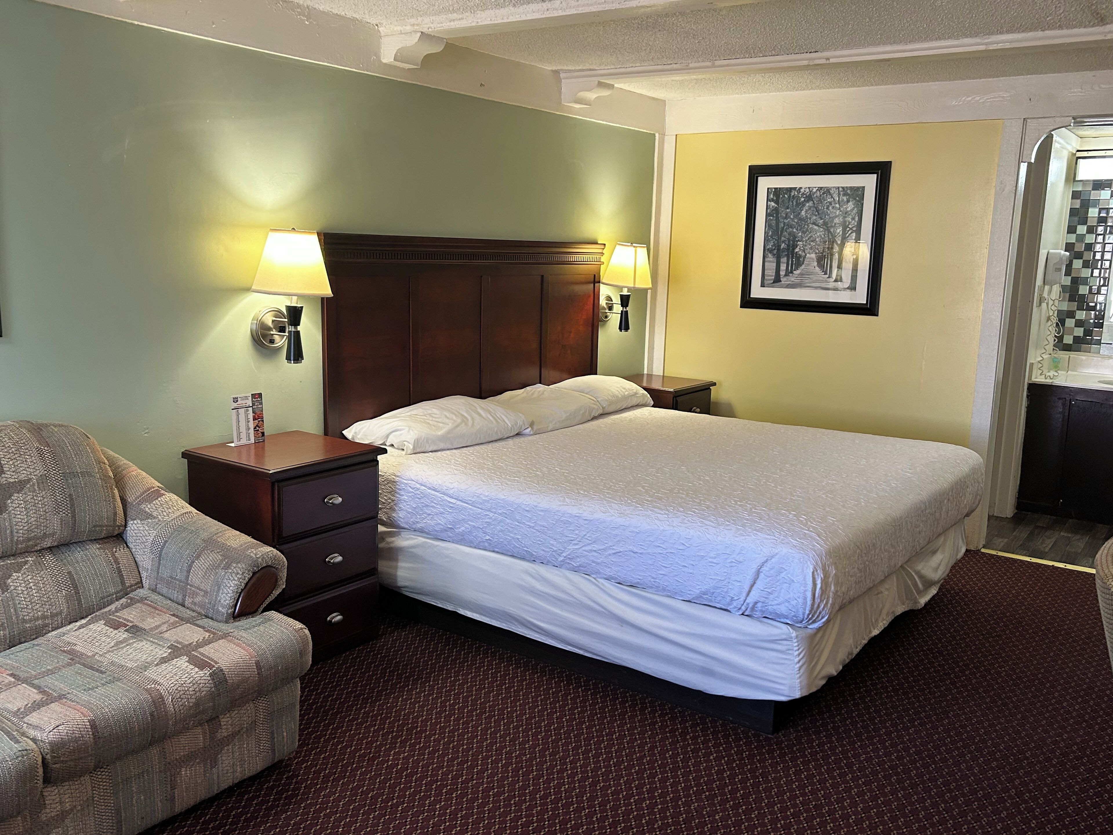 A hotel room with a large bed, two bedside tables with lamps, a wooden headboard, a patterned armchair, and a framed picture on the wall.