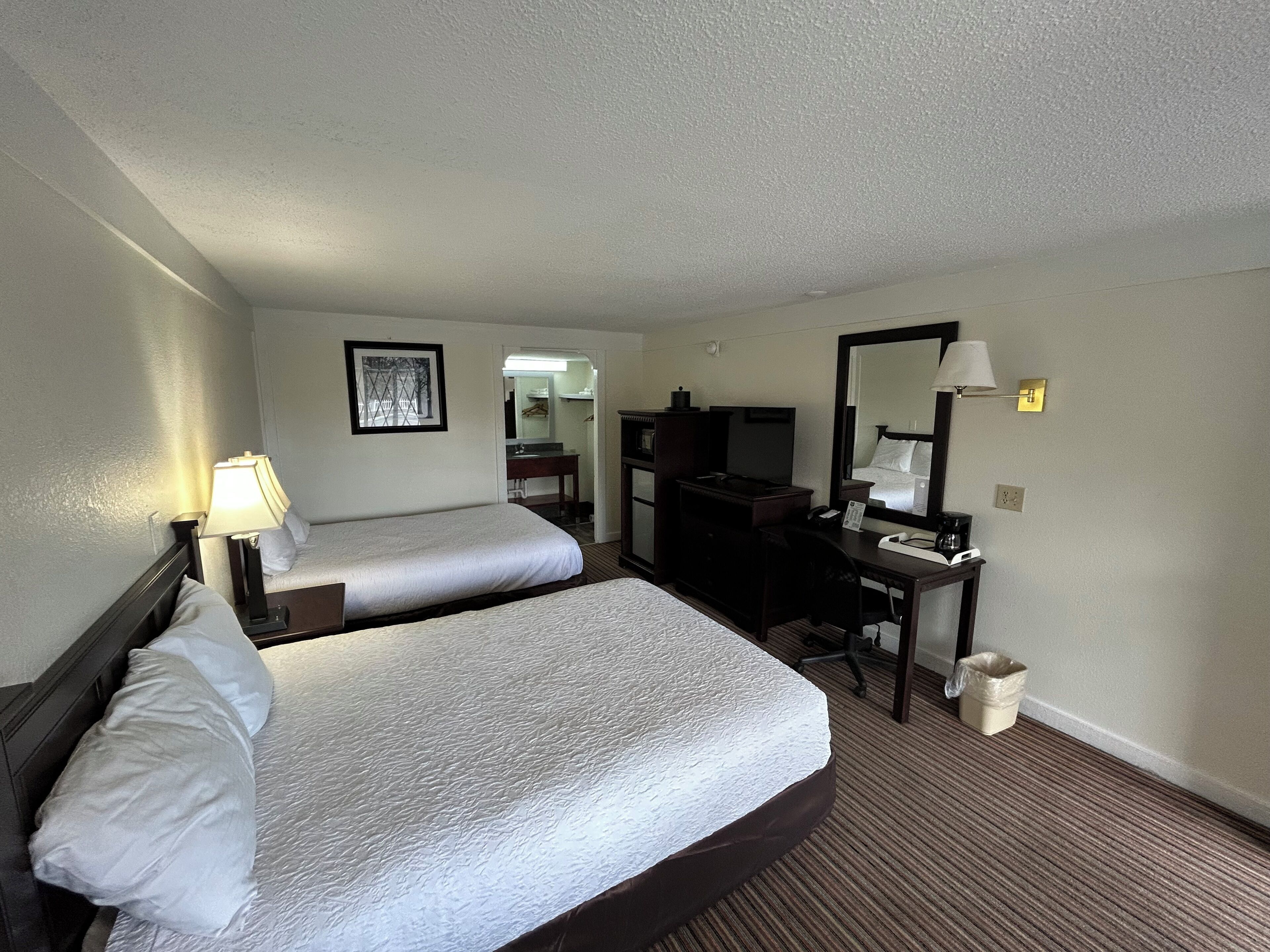 A hotel room with two beds, a desk, a chair, a mirror, and a lamp.
