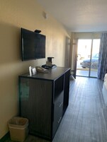 Habitación de hotel con un televisor de pantalla plana grande montado en la pared, un escritorio con teléfono y un cubo de basura pequeño, y vista al estacionamiento a través de la puerta.