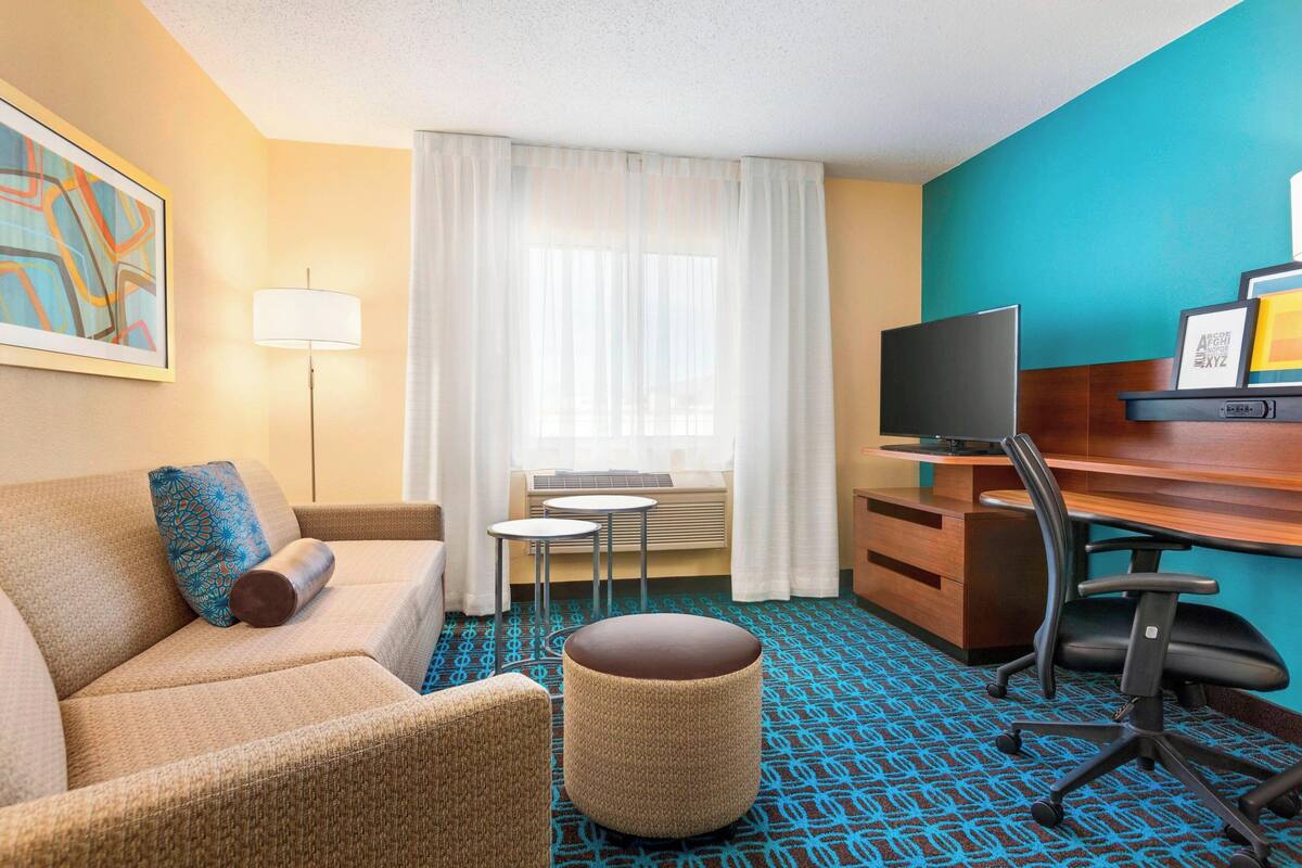A hotel room with a sofa, a small round ottoman, a desk with a chair, a television, and a window with curtains.