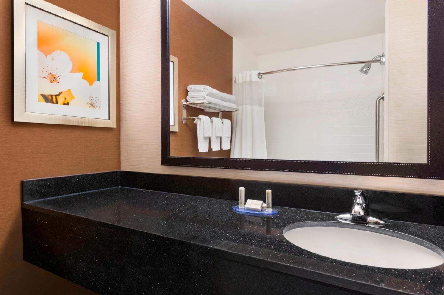 Un baño de hotel con encimera de mármol negro, lavamanos blanco, espejo y un cuadro en la pared.