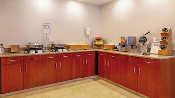 Uma área de cozinha com serviço de buffet, incluindo máquina de waffles, potes com frutas e um espaço para café.