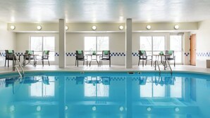 Una piscina interior con agua cristalina, rodeada de sillas y mesas, y grandes ventanales que permiten la entrada de luz natural.