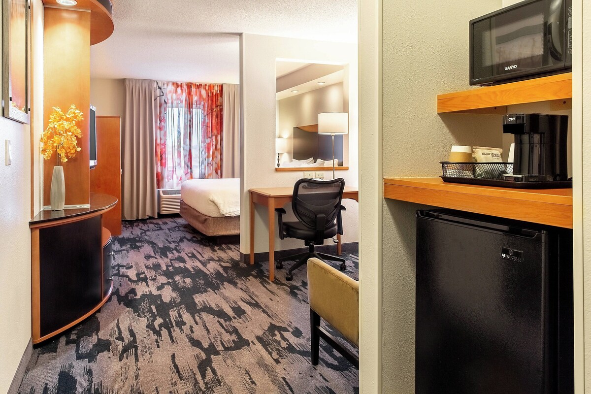 Una habitación de hotel con cama, escritorio, silla, minibar y un refrigerador pequeño.