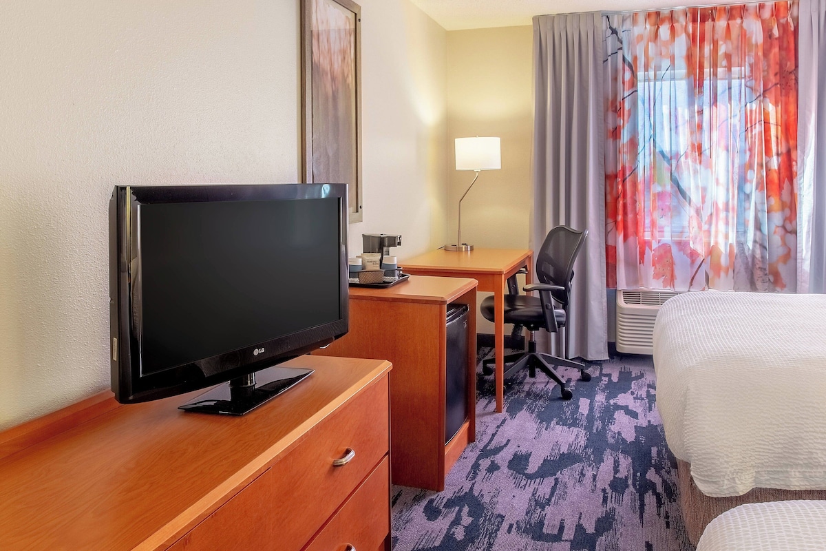 Habitación de hotel con televisión de pantalla plana sobre un aparador de madera, un escritorio con silla, una cama con colcha blanca y una ventana con cortinas florales.