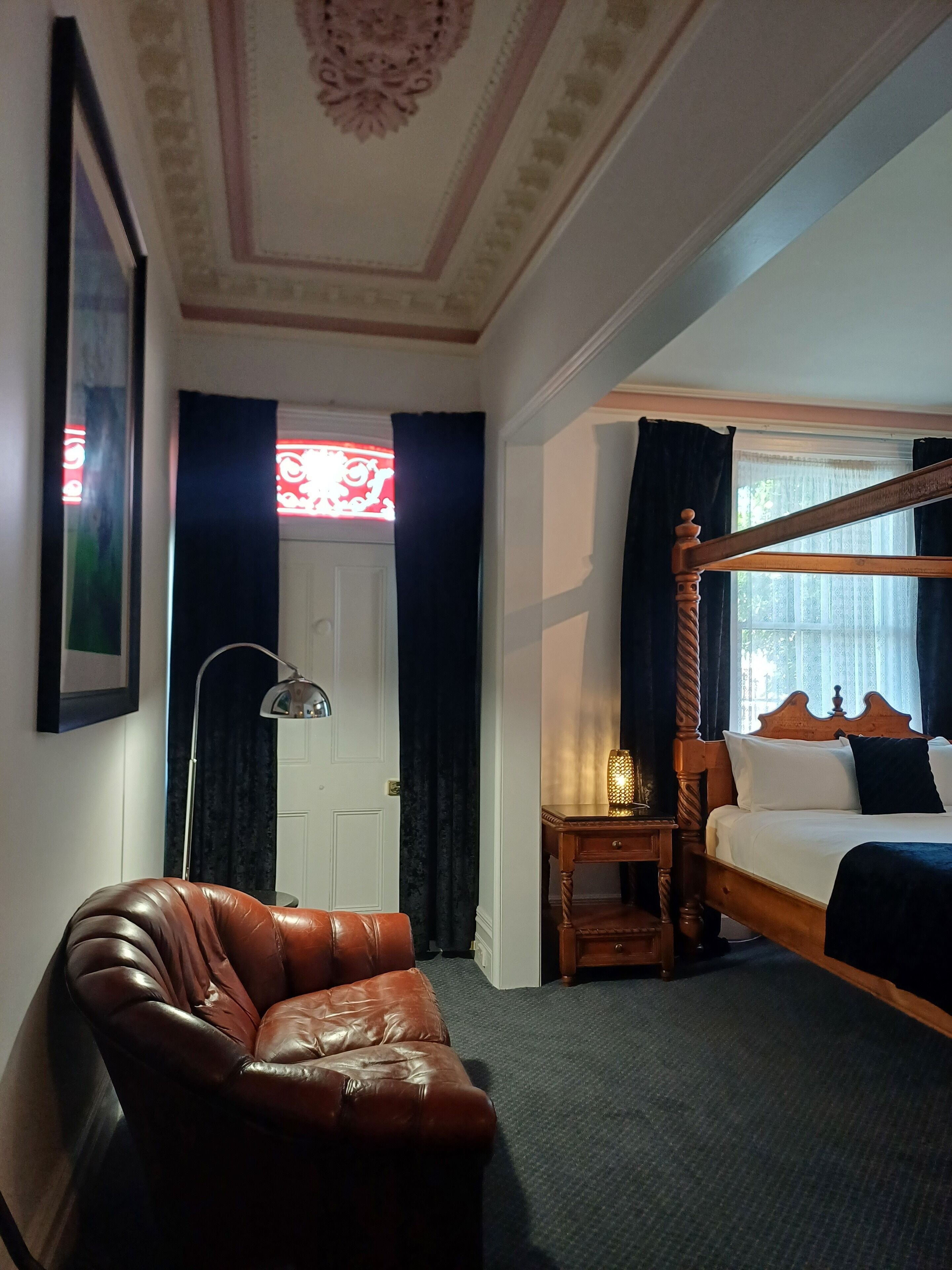 A room with a four-poster bed, a leather armchair, a desk with a lamp, and a framed picture on the wall.