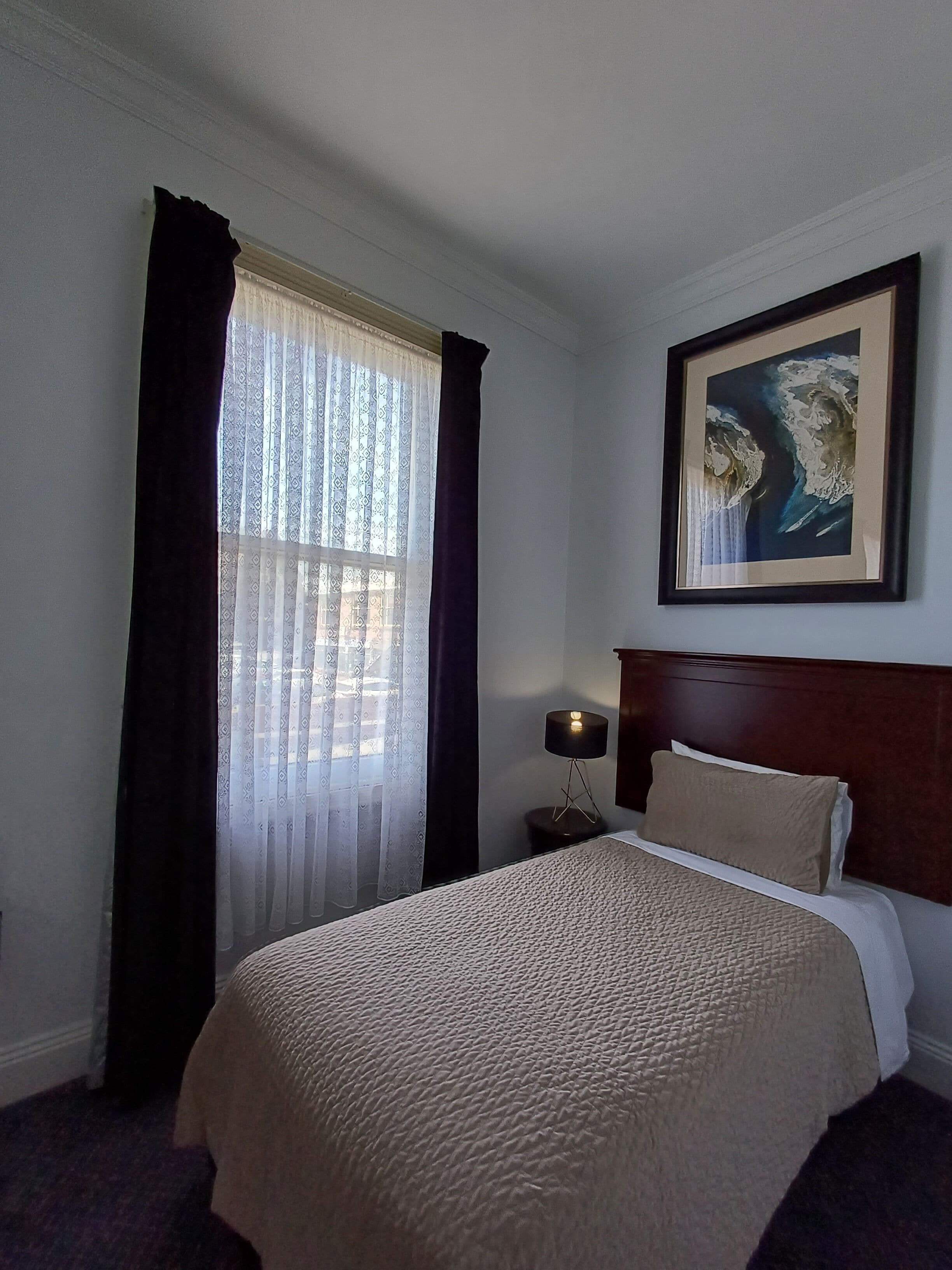 A hotel room with a bed, a nightstand with a lamp, a window with curtains, and a framed picture on the wall.