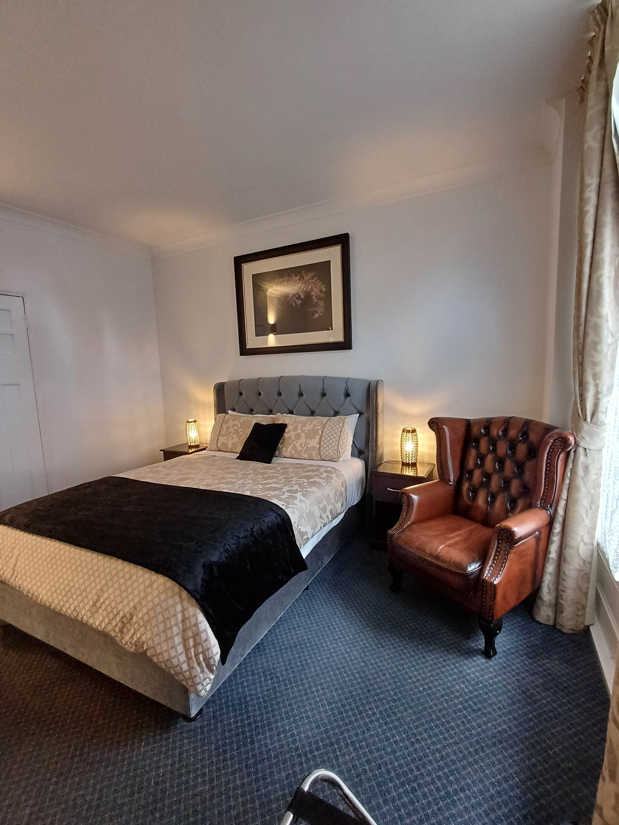 A hotel room with a large bed, a leather armchair, a framed picture on the wall, and a window with curtains.