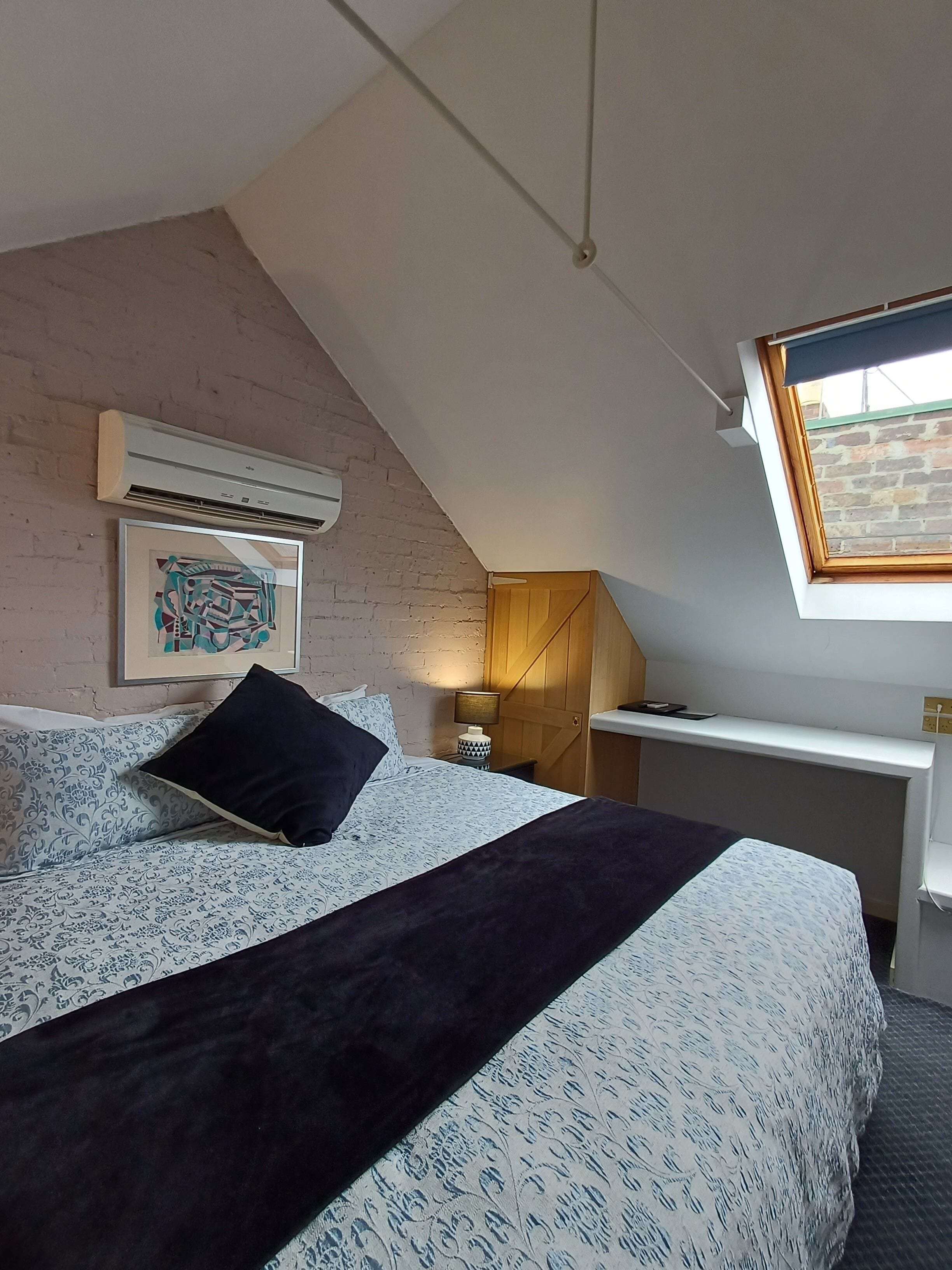 A bedroom with a sloped ceiling, a bed with patterned bedding, a wall-mounted air conditioner, a framed picture, a bedside lamp, and a window with a view of a brick wall.