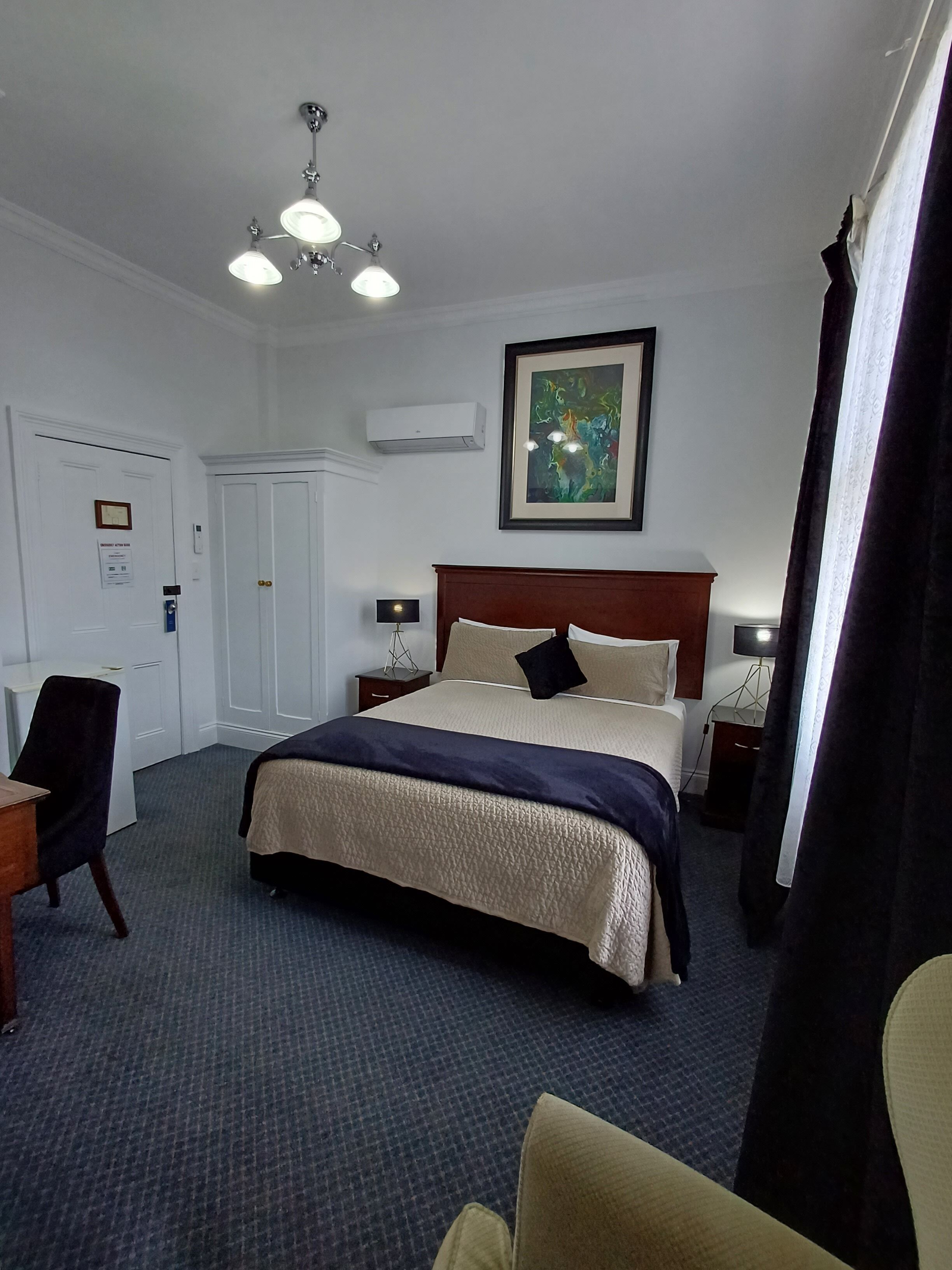 A hotel room with a large bed, a desk, a chair, a framed artwork on the wall, and an air conditioning unit.