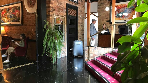 Un hall con pared de ladrillo, una planta grande, una silla roja, suelo de baldosas negras y una escalera con escalones rosados.