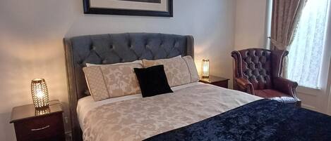 Un dormitorio ordenado con una cama grande, una silla, una mesita de noche y un cuadro en la pared.