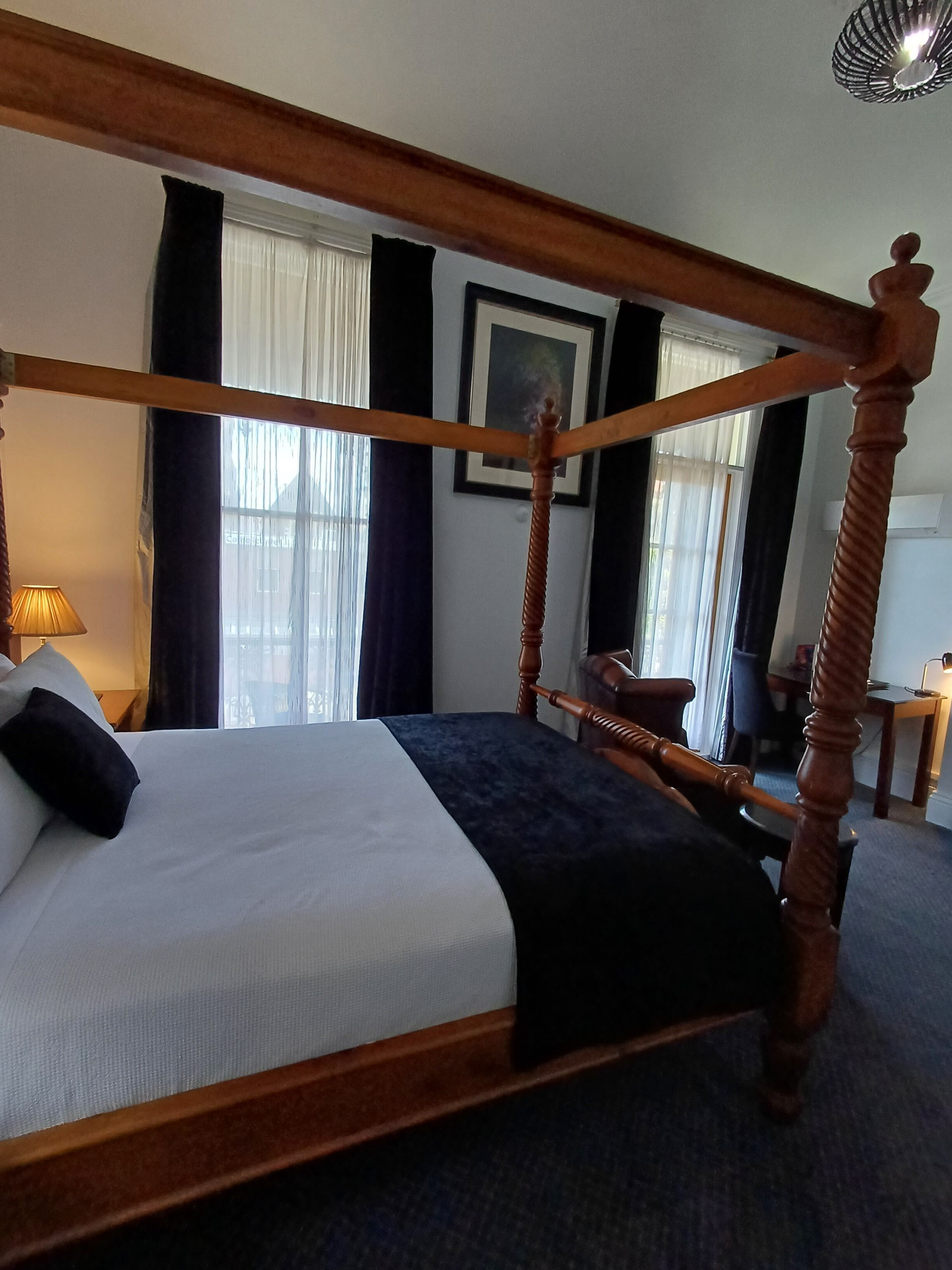 A four-poster bed with a dark headboard, white bedding, and a dark throw blanket. There is a lamp on a bedside table, a chair, and a desk with a computer in the room.