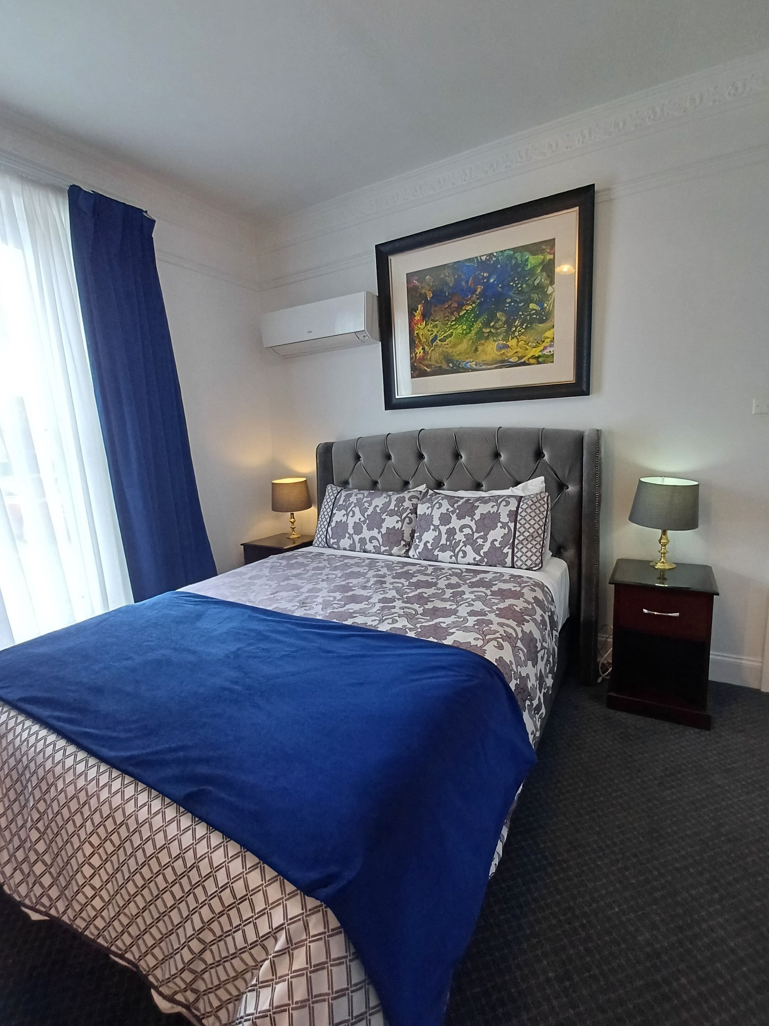 A hotel room with a large bed, a framed painting above the bed, a window with blue curtains, and a nightstand with a lamp.